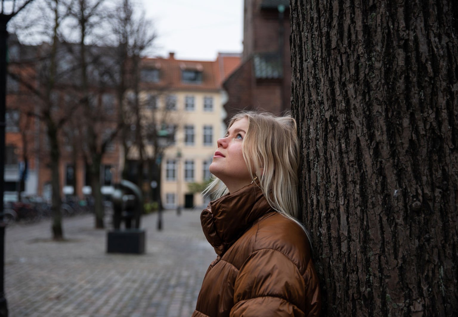 Ung tjej har en dunjacka och lutar sig mot ett träd och blickar upp mot himlen