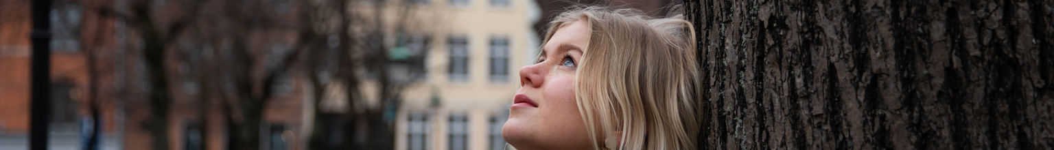 Ung tjej har en dunjacka och lutar sig mot ett träd och blickar upp mot himlen