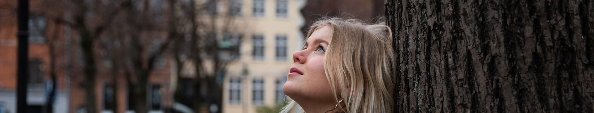 Ung tjej har en dunjacka och lutar sig mot ett träd och blickar upp mot himlen