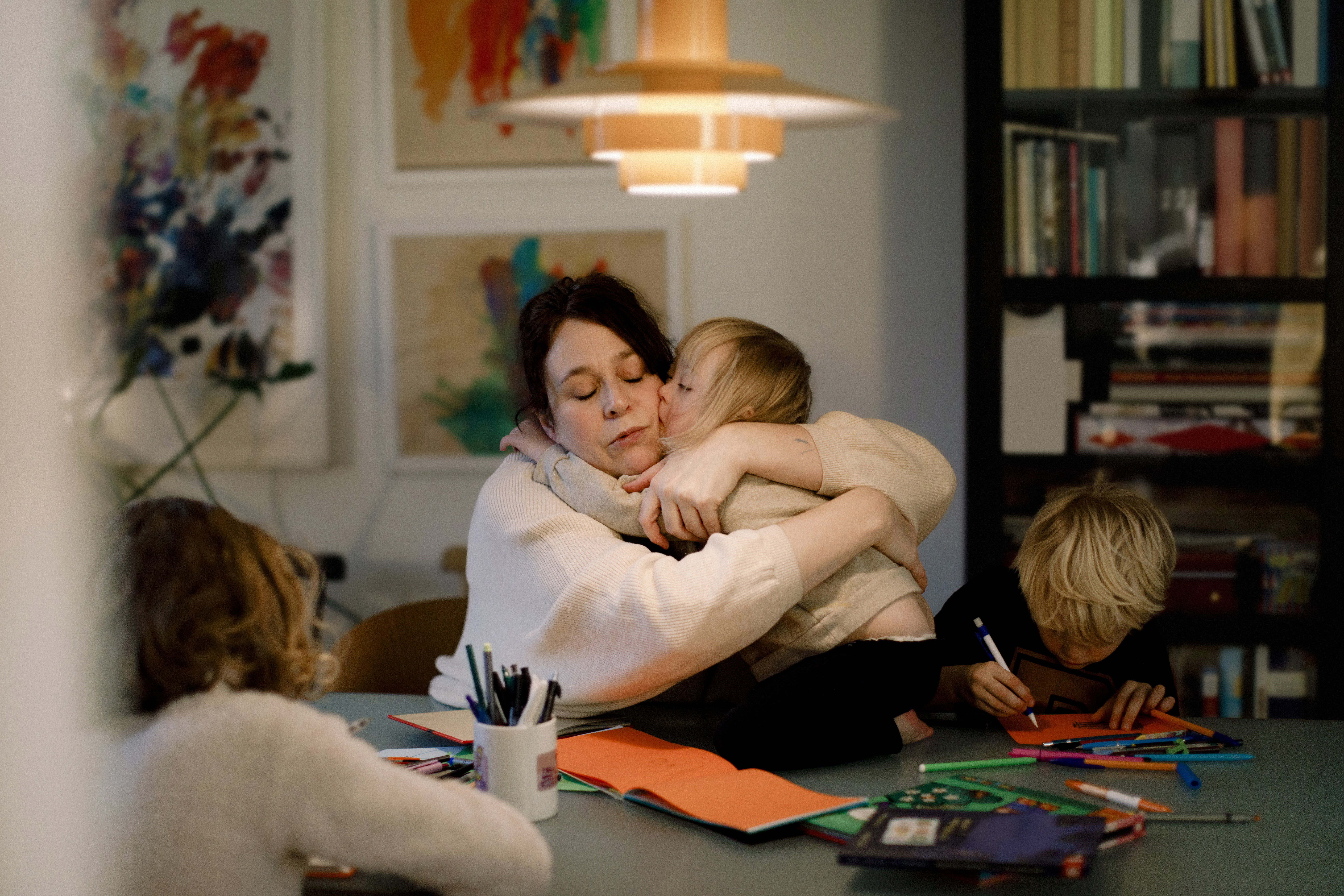 Mamma kramar om sitt barn vid ett bord där övriga pysslar med papper och penna