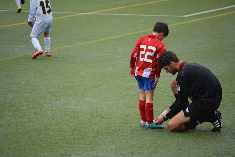 Ett barn får hjälp av fotbollsdomaren att knyta sina fotbollsskor på planen.