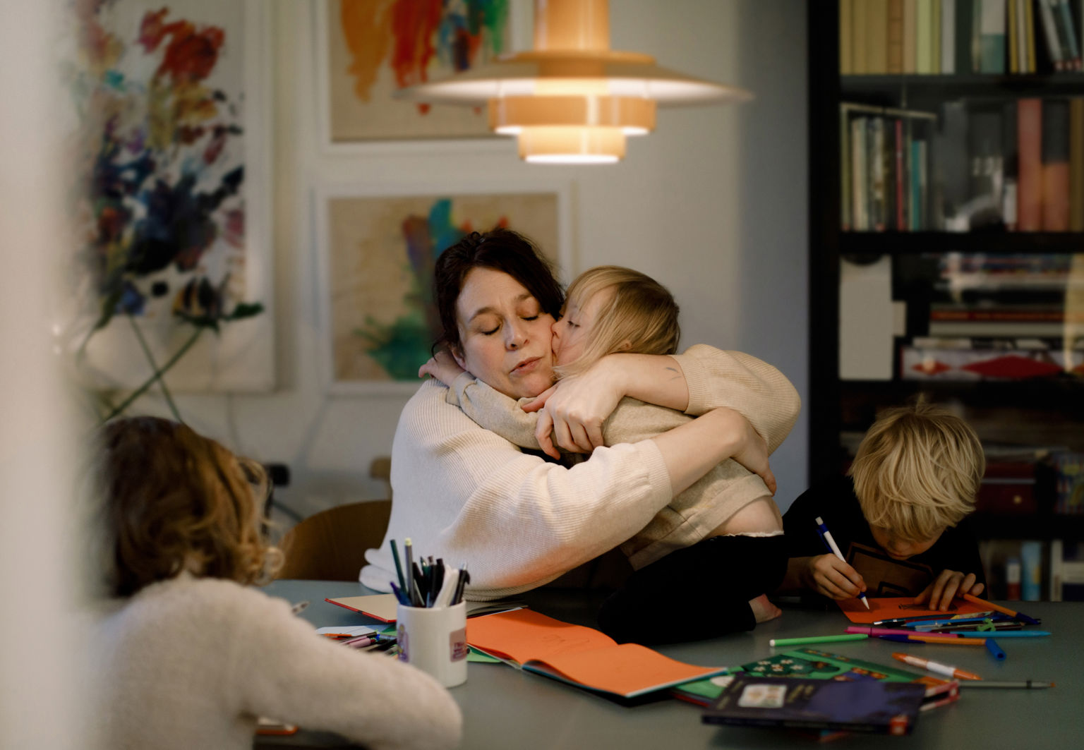 Mamma kramar om sitt barn vid ett bord där övriga pysslar med papper och penna